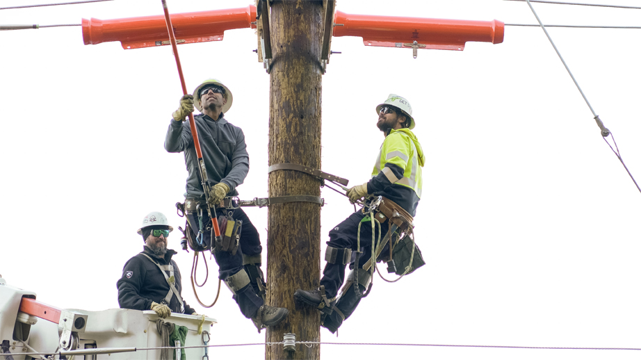 two linemen working on a utility pole wearing climbing gear with the lineman on the left holding out a hotstick and third lineman next to them in the bucket.